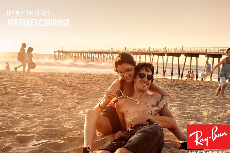 Ray-Ban sunglasses, a couple wearing the designer frames, sitting on the beach.